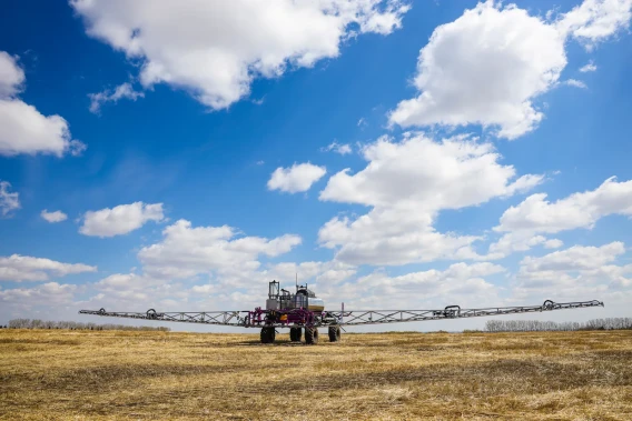 Agricultural field sprayer parked in an open field under a blue sky with clouds.