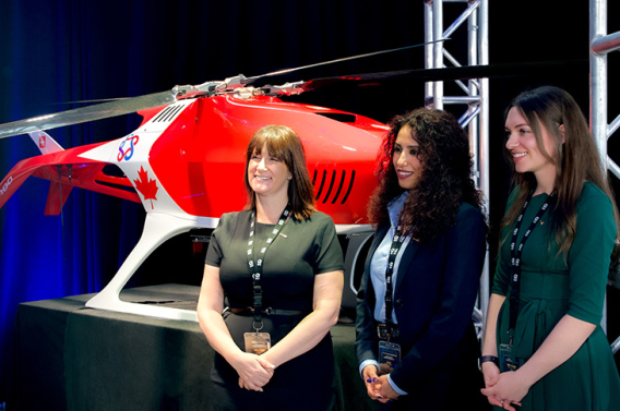 Three women stand beside a red drone or helicopter display at an event.