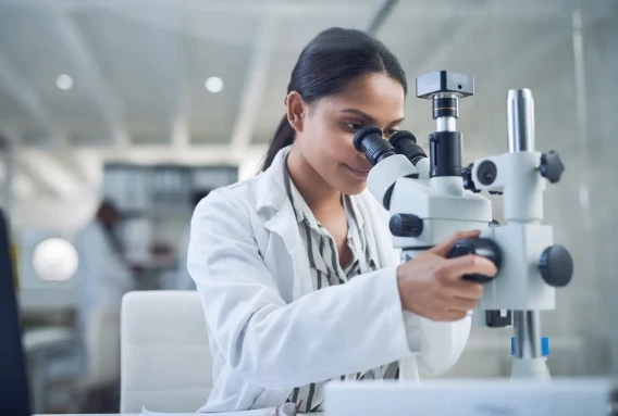 Scientist in a white lab coat adjusting and looking through a laboratory microscope.