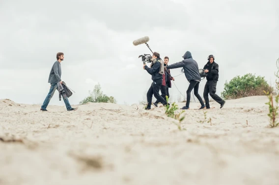 Film crew recording a tracking shot outdoors on a sandy landscape with camera, boom microphone, and sound team.