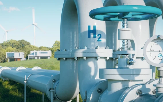 Close view of hydrogen pipeline infrastructure with valve, pressure gauge, wind turbines, and buildings in the background.