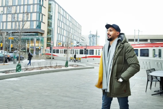 Man in a winter coat standing outside near a city train and modern buildings.
