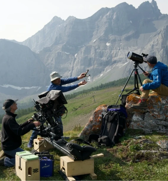 Film crew working on a mountain location with camera equipment, telescope, and clapperboard.