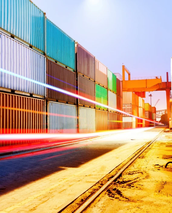 Rows of stacked shipping containers at a port with long light trails in the foreground at dusk.