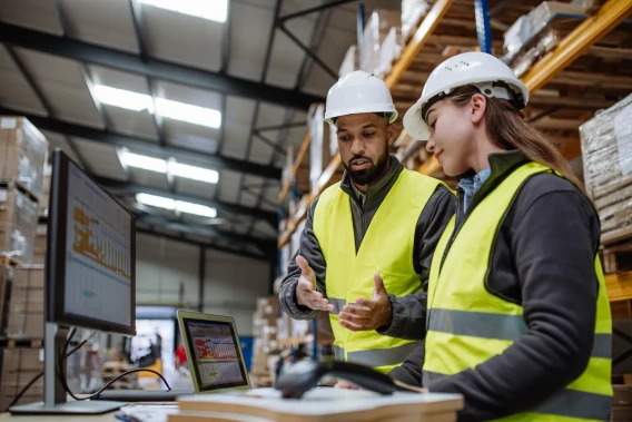 Two warehouse workers in safety vests and hard hats reviewing inventory data on a computer and tablet.
