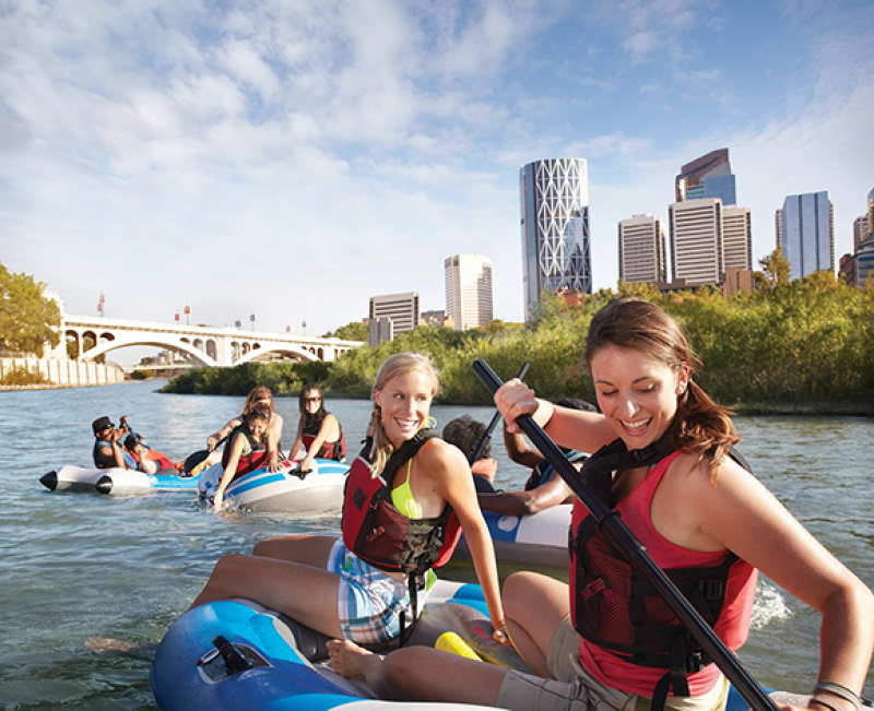 Group rafting on river with city skyline and bridge in background.