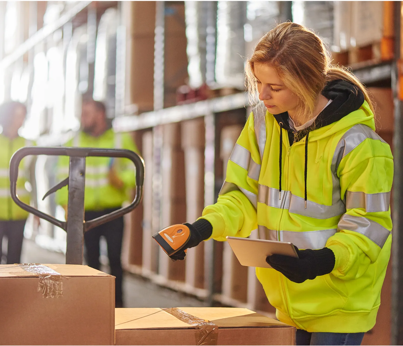 Warehouse worker in a high-visibility jacket scanning boxes with a handheld scanner and tablet.