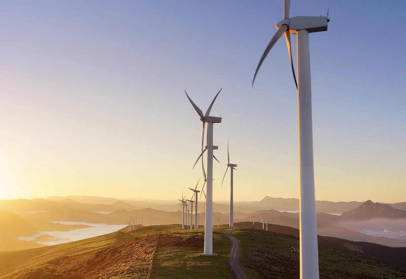 Wind turbines aligned along a hillside road during sunrise with mountains in the distance.